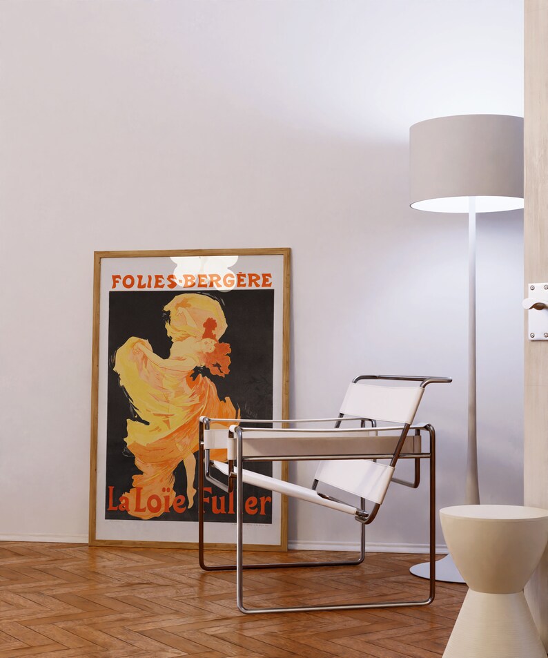 May include: A white leather and chrome chair sits in a room with a wood floor. A framed poster with the text "Folies-Berg&egrave;re La Lo&iuml;e Fuller" is on the wall behind the chair. A white floor lamp with a white shade is in the corner of the room.