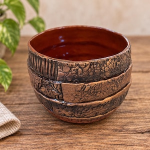 May include: A round ceramic bowl with a smooth, glossy reddish-brown interior and a textured, multi-paneled exterior featuring dark brown and copper-toned finishes with varied patterns including vertical lines and rough, organic rock-like surfaces.