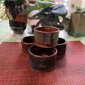 May include: Four brown ceramic cups stacked on top of each other. The cups have a dark brown glaze with a slight iridescent sheen. The cups are sitting on a red woven placemat.