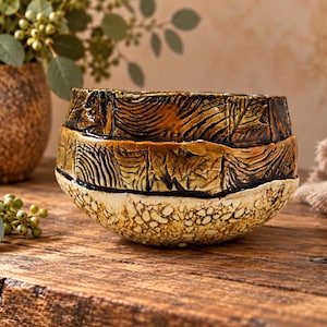 May include: A handcrafted ceramic bowl featuring a three-tiered design. The top section has a dark brown glaze with linear textures, the middle band shows a lighter brown finish, and the base has a cream-colored, speckled, textured surface.