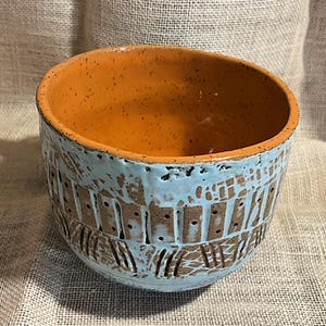 May include: Handmade ceramic bowl with a light blue exterior and a speckled orange interior. The bowl features a textured design with vertical and horizontal lines. A unique piece for serving or display.