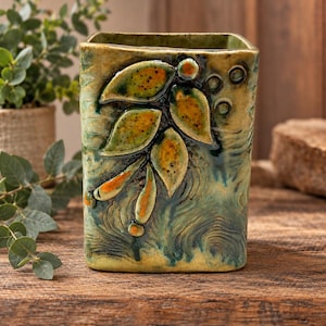 May include: A square ceramic vase with a glossy finish in shades of green, yellow, and brown. The front features a raised relief design of leaves and small circular accents, with a textured, swirling pattern across the surface.