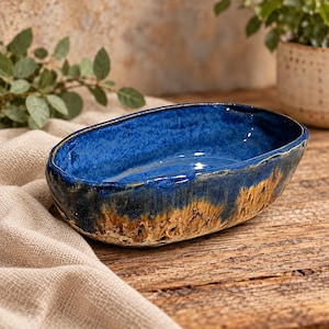 May include: An oval ceramic bowl featuring a deep blue glazed interior and a textured, earthy brown and tan exterior. The rim is uneven and organic in shape, showcasing a contrast between the smooth, glossy blue finish and the rough, mottled base.