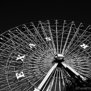 May include: A black and white photograph of a Ferris wheel with the word "TEXAS" spelled out in large letters on the wheel. The Ferris wheel is set against a dark sky.