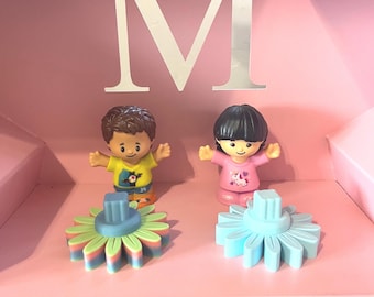 May include: Two small plastic figurines standing in front of a large letter M. One figurine wears a yellow shirt with a soccer ball graphic, the other a pink shirt with a unicorn. Two flower-shaped plastic bases in pastel colours sit in front.