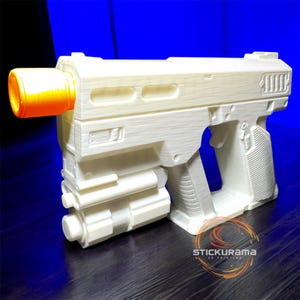 Peut inclure: Un pistolet jouet blanc imprimé en 3D avec un canon orange. Le pistolet jouet présente un design détaillé avec une surface texturée et une poignée. L'image est placée sur un fond bleu et une surface en bois sombre.