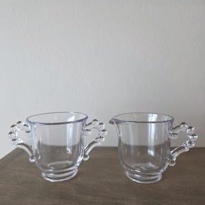 May include: Two clear glass serving pieces. One is a sugar bowl with two handles, and the other is a creamer with a spout. Both have decorative handles and a curved base. The items are on a brown surface.