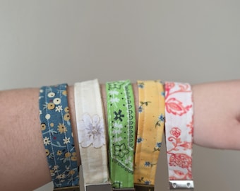 May include: A collection of five fabric wrist keychains with various floral and patterned designs. The keychains feature a blue floral, white flower, green paisley, yellow floral, and red floral pattern. Each keychain has a metal clasp and ring.