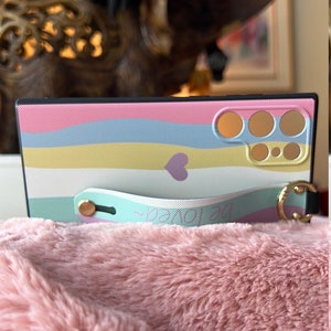 May include: A pink, blue, yellow and white striped phone case with a small purple heart and a gold ring. The case has a white strap with the words "Be Loved" written on it.