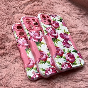 May include: Three pink and white floral phone cases with a raised camera lens design. The cases have a white background with pink and red floral patterns.