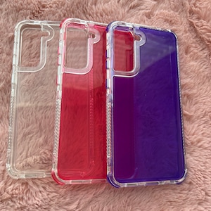 May include: Three clear phone cases with colored backs. The cases are lined up from left to right: clear, pink, and purple.