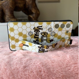 May include: A white phone case with a honeycomb pattern and gold accents. The case has a strap with the text "Bee Yourself" and a bee graphic.