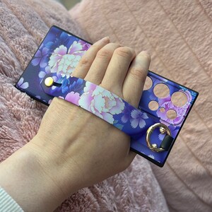 May include: A purple phone case with a floral design and a wrist strap. The case has cutouts for the camera and buttons.