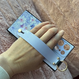 May include: A black phone case with a floral design and a light blue wrist strap. The case has a ring attached to it.