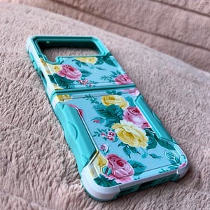 May include: A turquoise and white phone case with a floral pattern. The case has a raised edge around the camera lens and a cutout for the charging port.