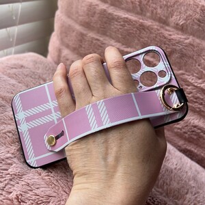 Könnte beinhalten: Rosa-weiß kariertes Handy-Case mit einem rosa-weißen Band. Das Band hat einen goldenen Metallring und einen schwarzen Knopf.