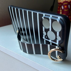 May include: Black and white striped phone case with a black strap and gold ring. The case has a cutout for the camera lens.