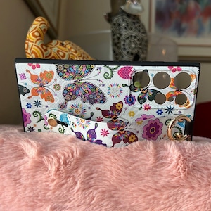 May include: A white phone case with a colorful butterfly pattern and a black strap. The case has a cutout for the camera lens and a gold ring on the strap.