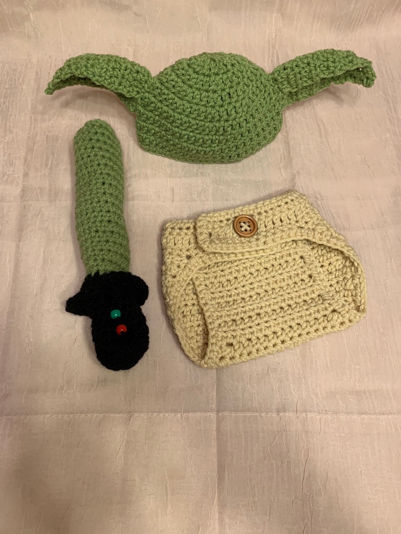 Puede incluir: Un disfraz de Yoda tejido a crochet para un beb&eacute;, que incluye un gorro, una cubierta de pa&ntilde;al y un sable de luz. El gorro es verde, la cubierta de pa&ntilde;al es beige y el sable de luz es verde y negro.