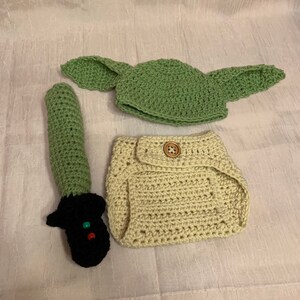 Puede incluir: Un conjunto de disfraz de beb&eacute; tejido a crochet que incluye un gorro verde con orejas, un pa&ntilde;al beige y un accesorio de sable de luz verde.