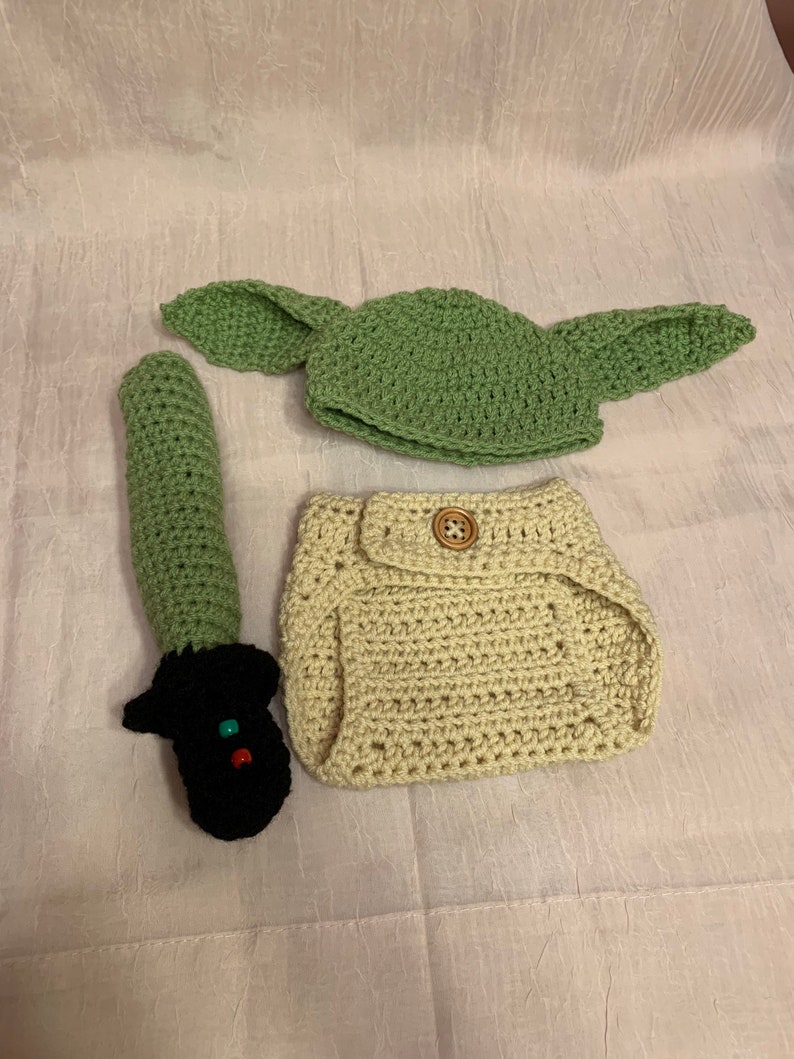 Puede incluir: Un conjunto de disfraz de beb&eacute; tejido a crochet que incluye un gorro verde con orejas, un pa&ntilde;al beige y un accesorio de sable de luz verde.