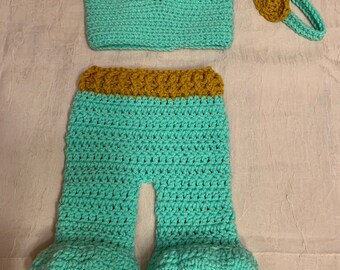 May include: A crocheted baby outfit in teal and gold. The outfit includes a top, pants, and a headband. The top has a V-neck and a gold trim. The pants have a gold waistband and are flared at the bottom. The headband is a simple band with a gold flower.