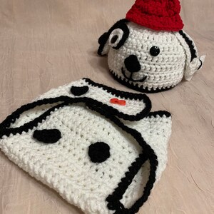 May include: White crocheted baby outfit with black trim and red buttons, featuring a dog face design. The outfit includes a matching hat with a red crocheted top.