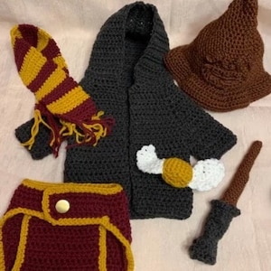 May include: A crocheted baby outfit inspired by the Harry Potter series. The set includes a hooded robe, a scarf, a hat, a nappy cover, and a wand. The robe is grey, the scarf is red and yellow, the hat is brown, the nappy cover is burgundy and yellow, and the wand is grey.