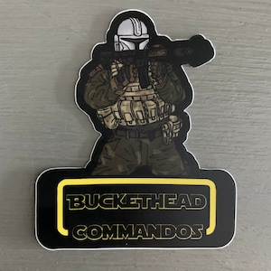 May include: A black and yellow sticker with a cartoon illustration of a person wearing a white helmet and camouflage clothing holding a rifle. The text "BUCKETHEAD COMMANDOS" is printed in yellow on the sticker.