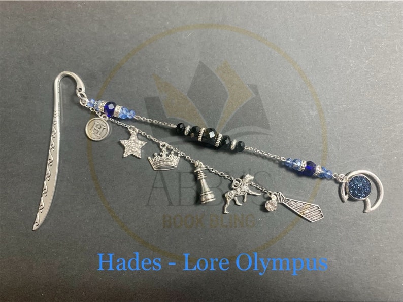 May include: Silver bookmark with a blue and black beaded chain and charms, including a crown, a star, a chess piece, a horse, a tie, and a letter H. The bookmark is labeled "Hades - Lore Olympus".