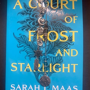 May include: A blue book with the title "A Court of Frost and Starlight" by Sarah J. Maas, a #1 New York Times bestselling author. A silver bookmark with charms hangs from the book.