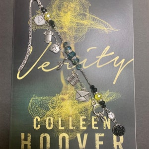 May include: A silver bookmark with a chain and charms, including a letter, a cross, a baby onesie, and a key. The bookmark is on top of a book titled "Verity" by Colleen Hoover, a #1 New York Times bestselling author.