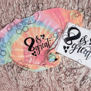 Könnte beinhalten: Zwei Batik-Shirts mit dem Text "8 is great" in schwarzer Schrift mit kleinen Herzen. Ein Shirt ist weiß und das andere ist ein Regenbogen-Batik.