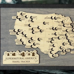 May include: Wooden map of the United States with cut-out locations and labels. The map is framed in black and includes a separate sign that reads "Supernatural America Travel Tracker". The map is made of wood.