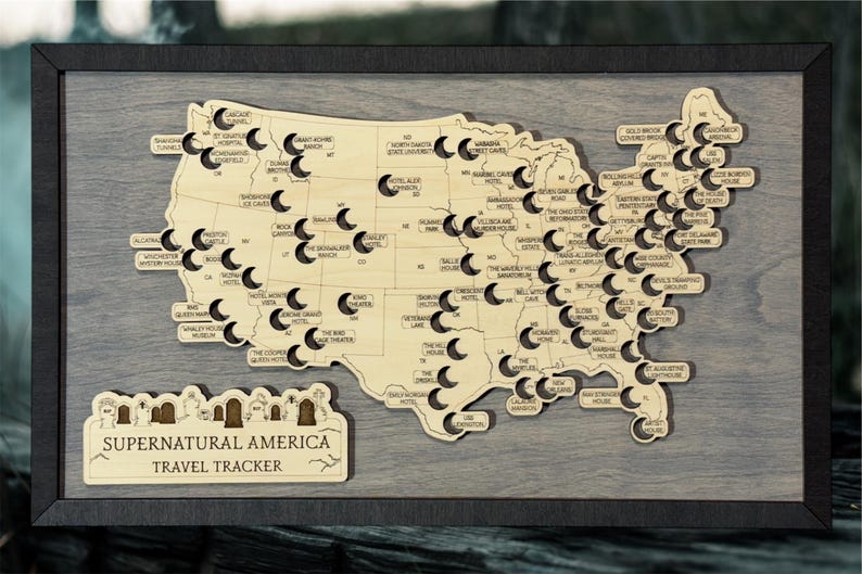 May include: Wooden map of the United States with cut-out locations and labels. The map is framed in black and includes a separate sign that reads "Supernatural America Travel Tracker". The map is made of wood.