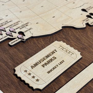 May include: Wooden map of the United States with laser-cut details, including amusement park icons and a ticket-shaped sign that reads "Amusement Parks Bucket List". The map is on a dark brown wooden surface.