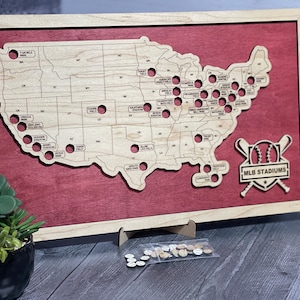 May include: Wooden map of the United States with cutouts for MLB stadiums. The map is set on a red background and framed in light wood. Includes small wooden markers and the text "MLB STADIUMS".