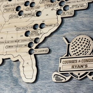 May include: A wooden golf course map of the United States with labeled golf courses. A separate wooden emblem features a golf ball, clubs, and the text "COURSES to CONQUER" and "RYAN'S". The map is on a blue surface.