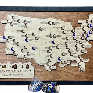 May include: Wooden map of the United States with cutouts and blue accents, framed in black. The map is labelled "Supernatural America Travel Tracker" and includes small, crescent-shaped markers. A small bag of markers is at the bottom.