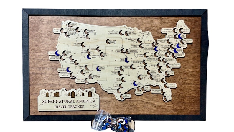 May include: Wooden map of the United States with cutouts and blue accents, framed in black. The map is labelled "Supernatural America Travel Tracker" and includes small, crescent-shaped markers. A small bag of markers is at the bottom.