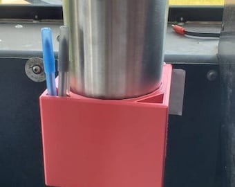 Peut inclure: Un porte-gobelet en plastique rouge avec un gobelet en acier inoxydable à l'intérieur. Deux stylos sont également dans le porte-gobelet. Le porte-gobelet est monté sur une surface noire.