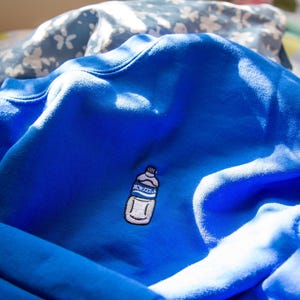 Może przedstawiać: Niebieska bluza z haftowanym białym obrazkiem butelki wody z napisem "AQUA ZOLA".