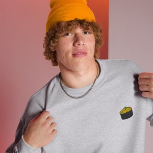 May include: A gray sweatshirt with a small embroidered image of a yellow and brown pastry on the left chest. The model is wearing a yellow beanie and a silver chain.
