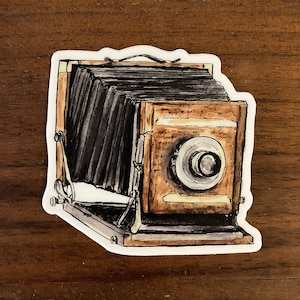 May include: A vintage-style camera sticker with a black bellows, a wooden body, and a lens with a circular shutter. The sticker has a white border.
