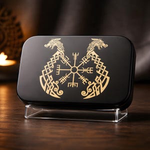 May include: A black rectangular box featuring a gold Viking symbol and dragon design. The box is presented on a clear stand, highlighting the detailed artwork. The background is blurred, drawing focus to the box's design and craftsmanship.