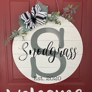 May include: A white round wooden door hanger with a gray letter "S" and the text "Snodgrass Est. 2020" in black. The door hanger is decorated with a black and white striped bow and green eucalyptus leaves. The word "welcome" is written in white on a red door.