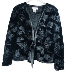 May include: A black velvet jacket with a floral pattern. The jacket has a tie closure and long sleeves. The floral design is a lighter shade of black, creating a subtle contrast. The jacket is hanging on a white hanger.