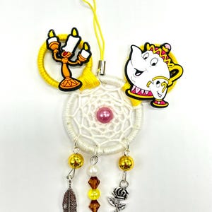 Puede incluir: Un atrapasueños blanco con detalles amarillos y dorados que presenta una tetera amarilla con una cara blanca y un candelabro amarillo con una cara blanca. El atrapasueños tiene una cuenta rosa en el centro y una pluma plateada, una rosa plateada y cuentas doradas y blancas que cuelgan de la parte inferior.