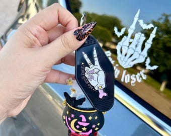 May include: A black motel-style keychain tag featuring a white hand making a peace sign with the text BAD WITCH VIBES, attached to a black cauldron-shaped accessory decorated with a pink crescent moon, stars, and a witch hat graphic.