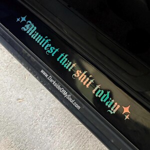 May include: A black car door with a sticker that reads "Manifest that shit today" with a website address below it. The sticker is in a gradient of teal and pink.